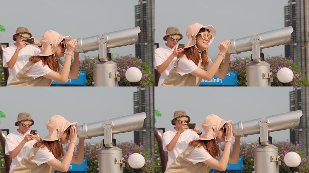 欢快的亚洲年轻夫妇游客在观看点使用投币式双筒望远镜观看城市背景。