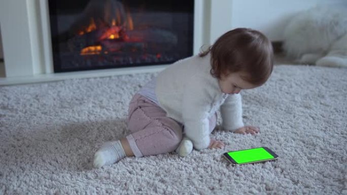 可爱开朗的幸运聪明的学步宝宝女孩在蓬松的地毯上使用智能手机绿屏手机Hromakey看视频卡通放松在家