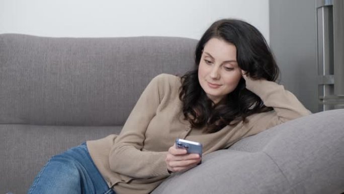女人躺在家里的沙发上，用手机滚动新闻。使用智能手机上的社交媒体应用程序的30年代美丽黑发模特的股票视