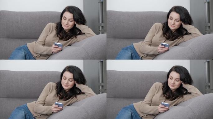女人躺在家里的沙发上，用手机滚动新闻。使用智能手机上的社交媒体应用程序的30年代美丽黑发模特的股票视