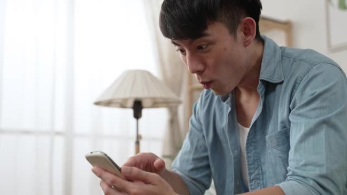 特写一位震惊的台湾男性在手机屏幕上张开嘴向下滚动，同时在家里的手机上阅读令人惊讶的在线新闻。