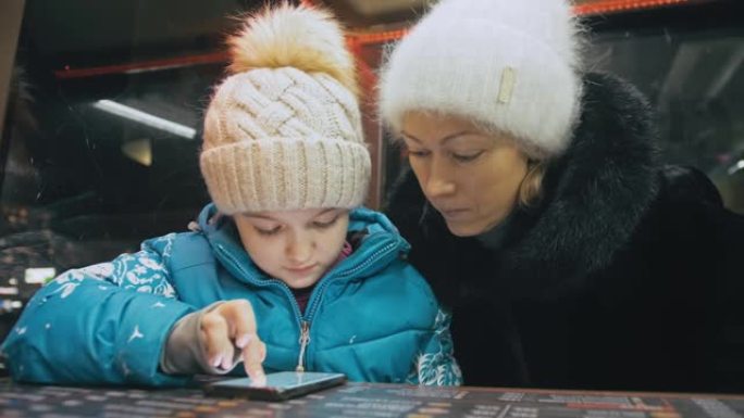 迷人的高加索母亲和女儿使用智能手机坐在咖啡馆。高级小孩在智能手机上向妈妈展示和解释一些东西