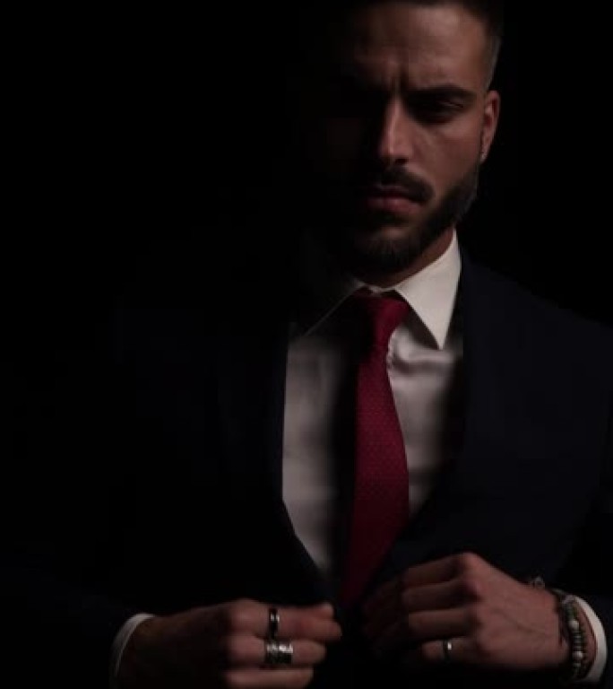 有吸引力的年轻商人扣扣西装，固定领带并向侧面看，在侧视图位置移动并在黑暗中褪色的垂直视频