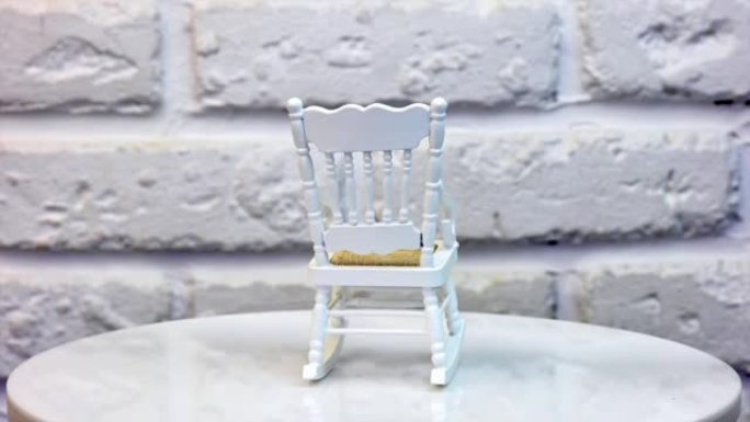 娃娃屋的白色小摇椅。旋转桌上的一件相当小的家具。背景上的白色砖墙。特写。