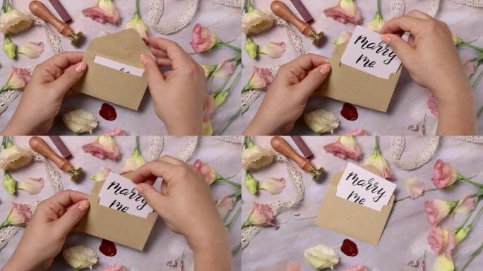 双手在粉红色的花朵附近打开带有MARRY ME卡的信封
