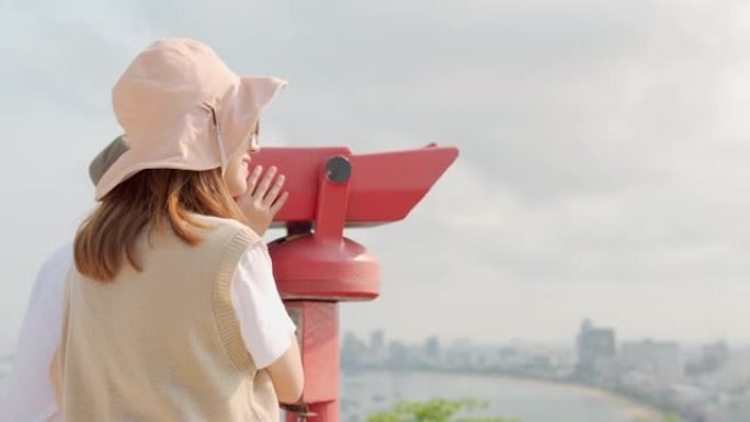 欢快的亚洲年轻夫妇游客在观看点使用投币式双筒望远镜观看城市背景