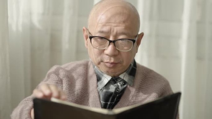 一位亚裔美国老人坐在窗边看书戴着老花镜