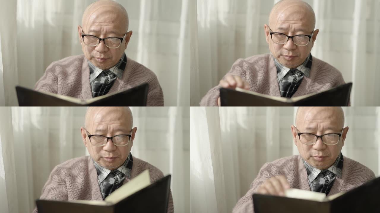 一位亚裔美国老人坐在窗边看书戴着老花镜