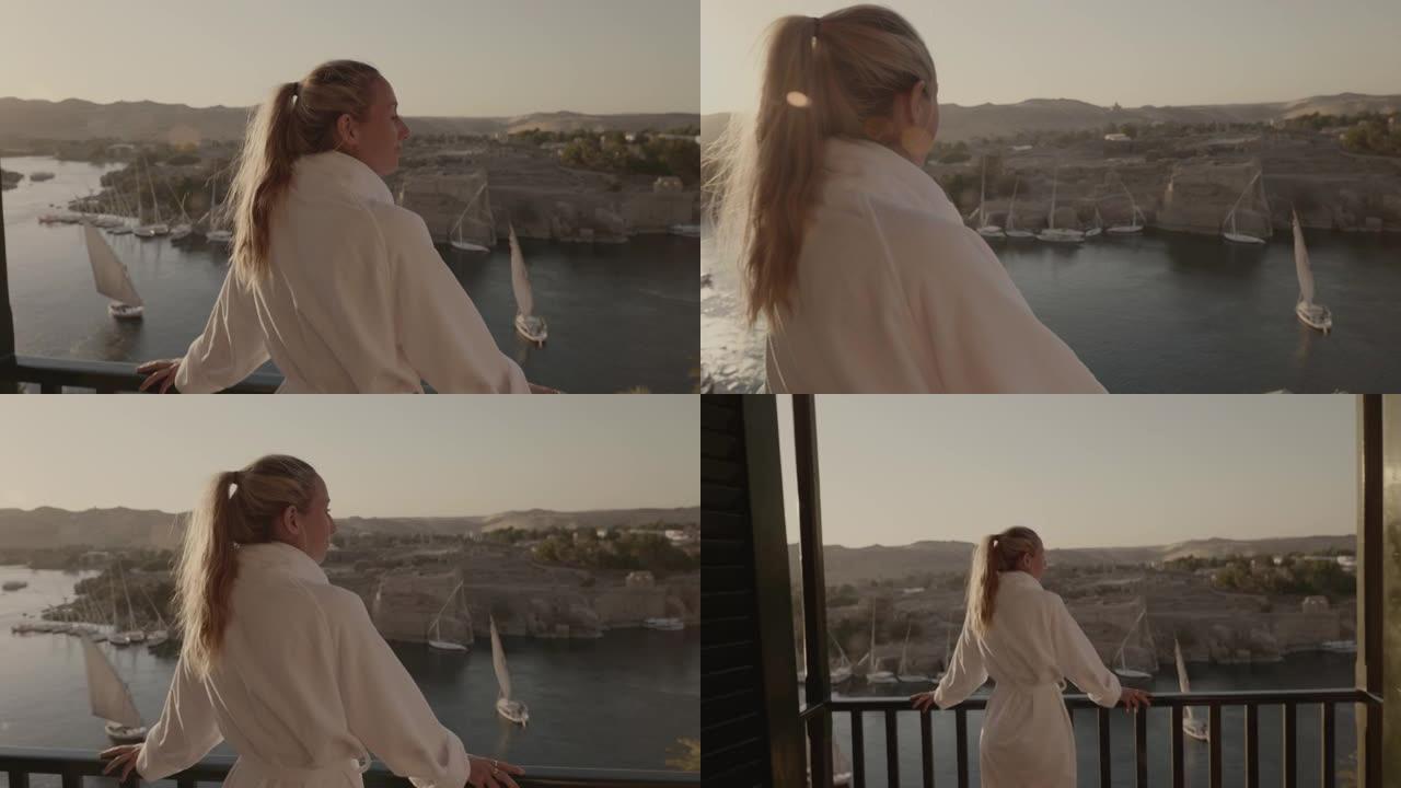 女人站在阳台上，在日落时欣赏壮丽的河景。日落时漂浮在尼罗河上的帆船。