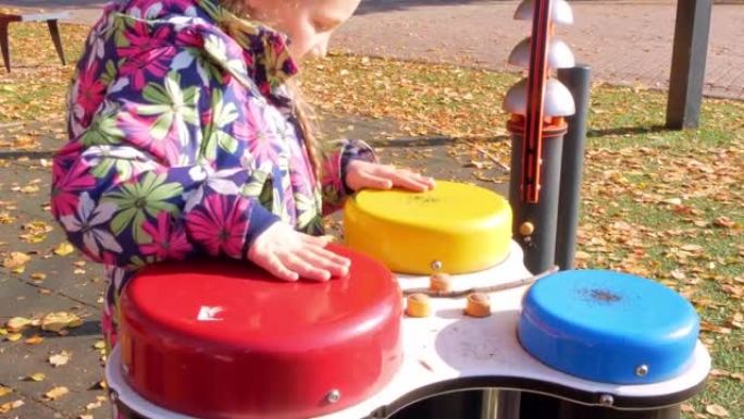 一个小女孩在秋天城市公园的操场上用手敲着邦戈鼓。孩子玩音乐与彩色彩虹儿童鼓设置在秋天的街道。可爱快乐