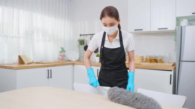 亚洲清洁服务女工在家厨房打扫卫生。漂亮的女管家清洁工戴口罩，用鸡毛掸子擦拭凌乱的柜台做家务或家务。