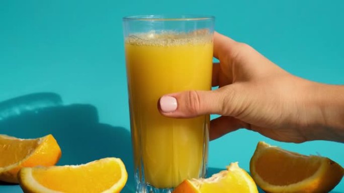 女性手放一杯新鲜的榨橙汁，切成薄片的水果在蓝色背景上。果汁，新鲜冷饮。夏天提神。水果切成两半