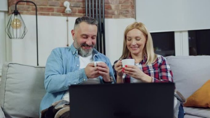 美丽幸福的爱心成年夫妇晚上坐在家里，在电脑上观看激动人心的电影时碰碰茶杯，家庭休闲概念