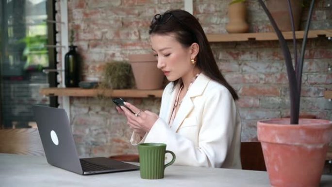 在咖啡馆餐厅办公室穿着经典服装的年轻中国亚洲女商人的肖像使用她的手机和笔记本电脑。自由职业者在咖啡店