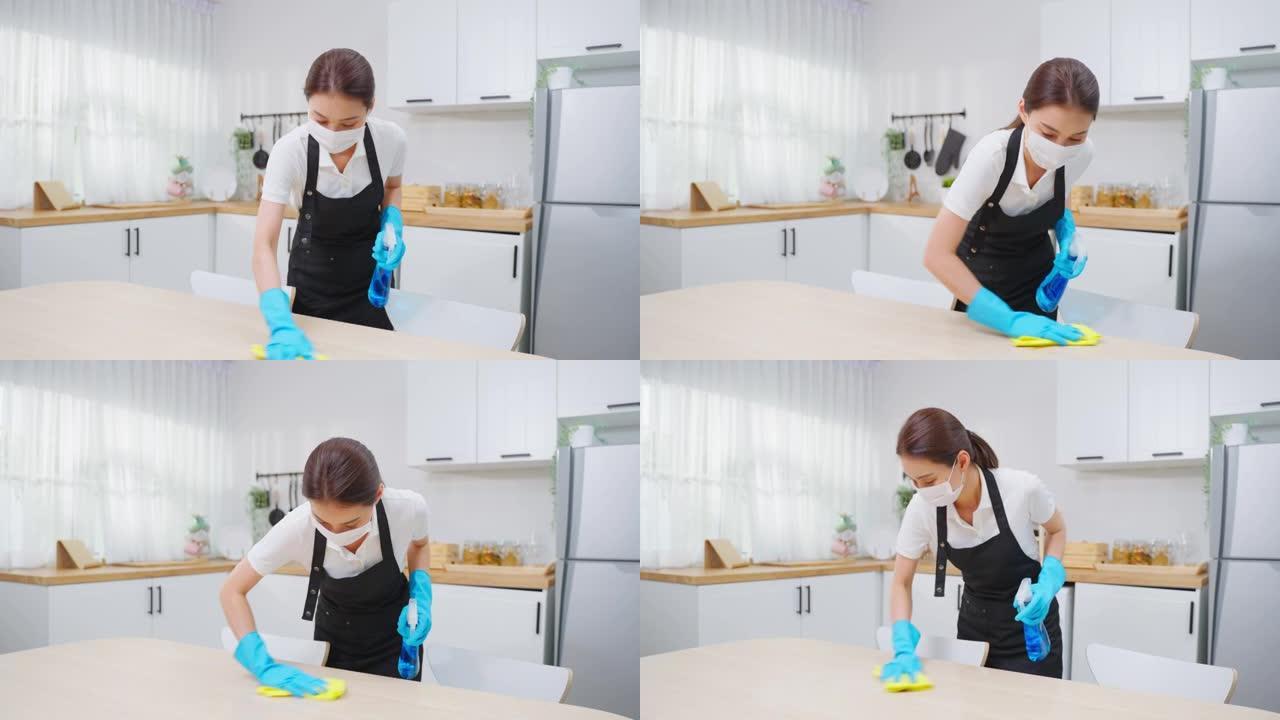 亚洲主动清洁服务女工在家厨房打扫卫生。漂亮的年轻女孩管家清洁工戴口罩，感到快乐，擦拭脏乱的脏桌子做家