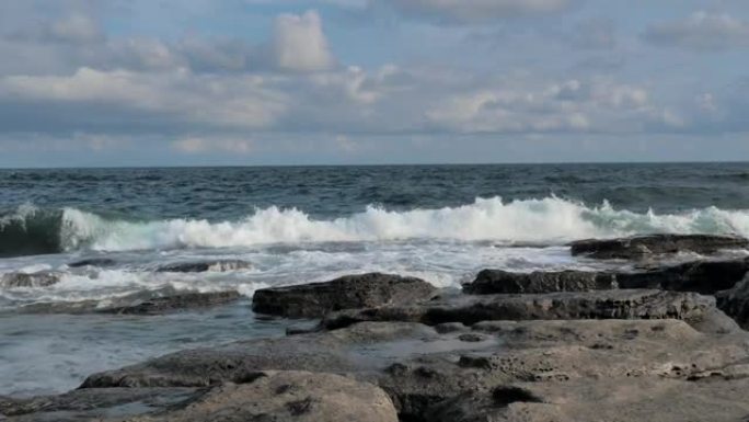 海浪溅落在蓝色海洋背景的岩石海岸上。波涛汹涌的岩石海岸线在暴风雨中席卷而来。海浪用泡沫在石头上破裂。