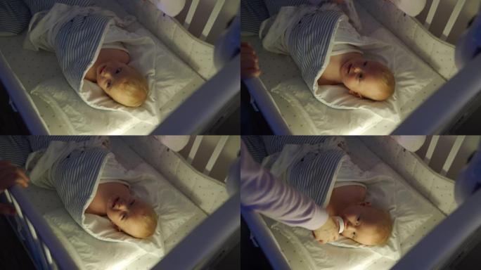 母亲襁褓中的婴儿睡在婴儿床里，面对襁褓中的9个月男婴在夜灯下。