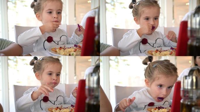 可爱的2岁小女孩，独自吃午饭，一边看平板电脑上的卡通片，一边在餐厅的家庭午餐时间吐蔬菜沙拉