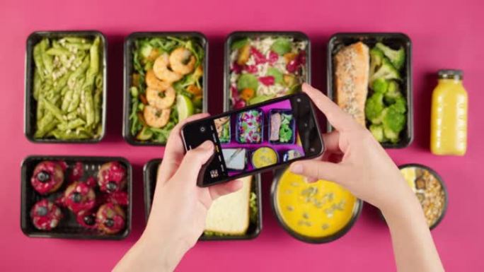 送餐顶视图，在粉红色背景的一次性容器中带走餐点。在智能手机上拍摄带有煮熟的素食菜肴的午餐盒照片。健康