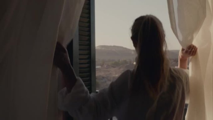 特写后景年轻女子打开窗帘站在阳台上的豪华酒店房间里，早上欣赏迷人的河景