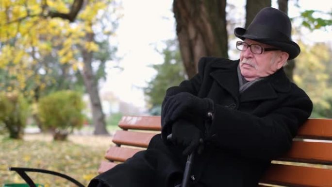 资深孤独的祖父在秋天的城市公园里用棍子坐在长凳上。