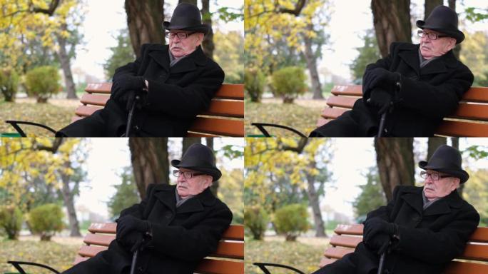 资深孤独的祖父在秋天的城市公园里用棍子坐在长凳上。