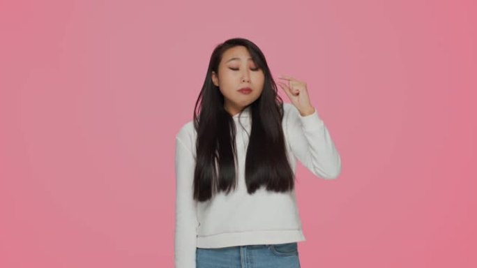 沮丧的年轻亚洲女孩用手指展示小尺寸手势，粉红色工作室背景上的小尺寸