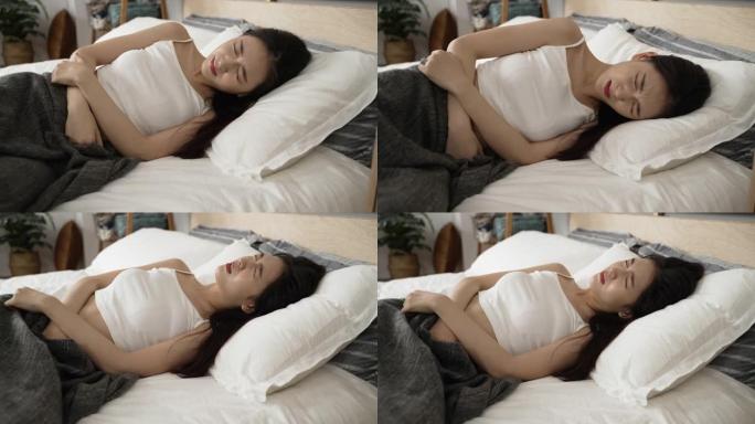 下腹部严重抽筋的亚洲女孩白天抱着肚子躺在床上，在家里的卧室里呻吟着。