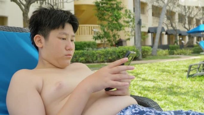 快乐的亚洲男孩使用智能手机，在酒店度假期间放松附近的游泳池，与家人、生活方式理念。