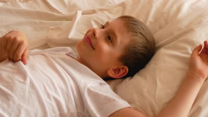 一个7-9岁的高加索男孩的特写镜头在白天醒来，心情很好。孩子醒来，躺在床上时伸出双臂。早上好，我醒了
