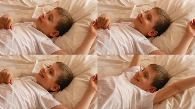 一个7-9岁的高加索男孩的特写镜头在白天醒来，心情很好。孩子醒来，躺在床上时伸出双臂。早上好，我醒了