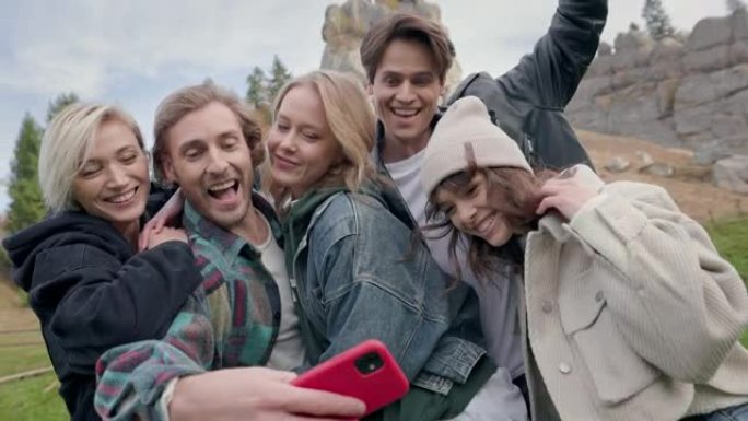大自然中的五个朋友自拍。两个年轻男人和三个女人在镜头前一起微笑。留着胡子的男人从口袋里掏出手机，其他