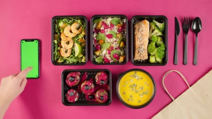 送餐顶视图，在粉红色背景的一次性容器中带走餐点。午餐盒配有煮熟的素食菜肴，使用带色度绿屏的手机。健康