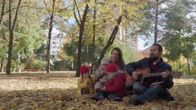 一个幸福的大家庭，有小孩，在公园野餐中度过时光，爸爸弹吉他唱着有趣的歌。看秋天公园的相机寒冷天气。童