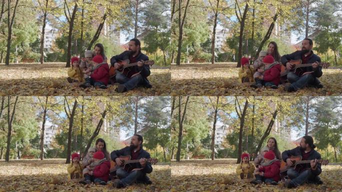 一个幸福的大家庭，有小孩，在公园野餐中度过时光，爸爸弹吉他唱着有趣的歌。看秋天公园的相机寒冷天气。童