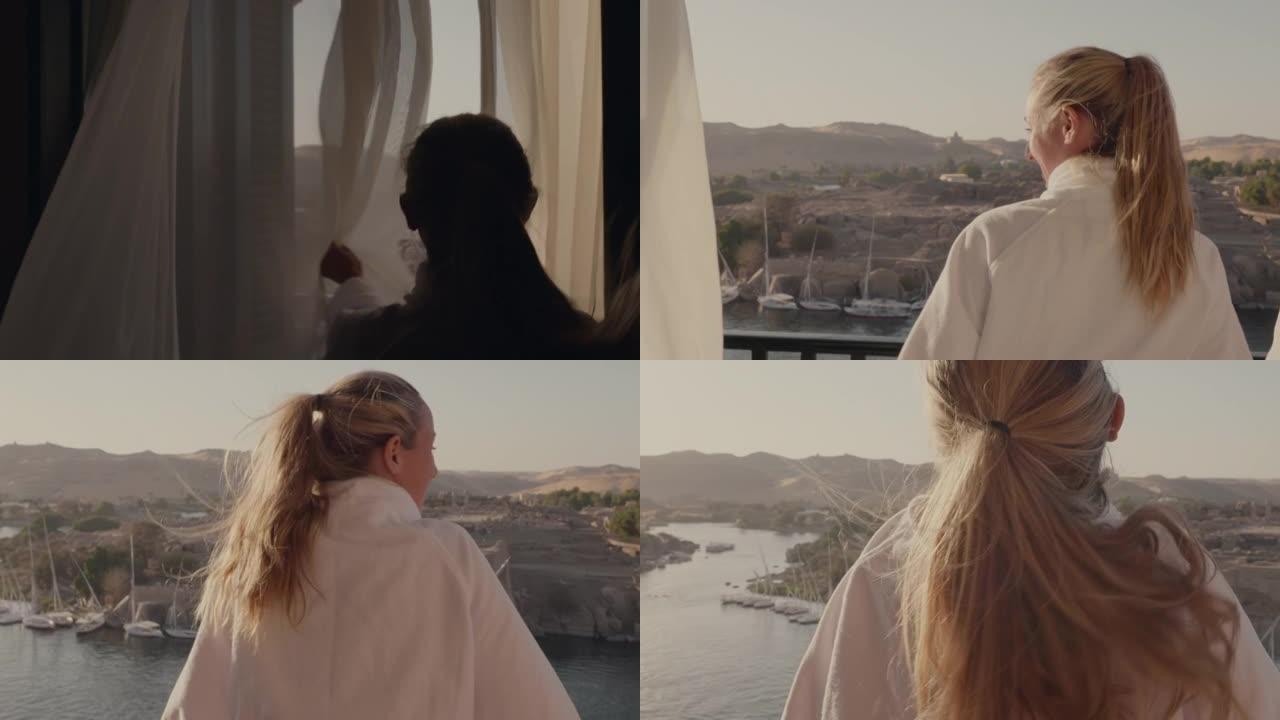 走向房间窗户的年轻女子打开窗帘，走到阳台上。埃及阿斯旺早上壮观的河景和帆船