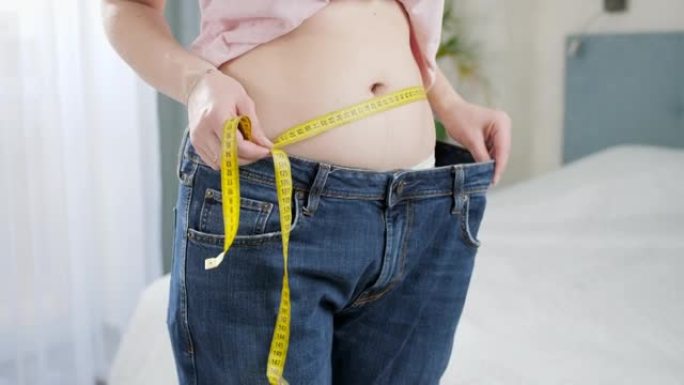 美丽的年轻女子，苗条的腹部用胶带测量腰部，在镜子里展示大牛仔裤。节食、减肥和健康的生活方式的概念。