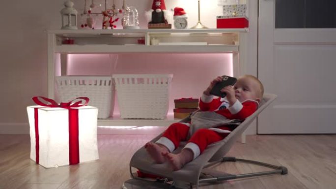 快乐的婴儿穿着圣诞老人服装，使用智能手机坐在房间里，带着圣诞节日的灯光，可爱的孩子庆祝他的第一个新年