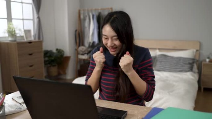 快乐的韩国女学生收到录取通知书电子邮件，握紧拳头，向前倾斜电脑屏幕，在家中查看细节。
