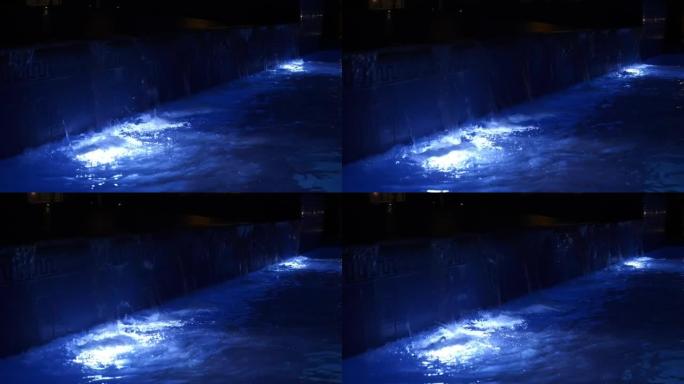 晚上的游泳池。特写。泳池水，蓝色背光，晚上。游泳池里的小瀑布