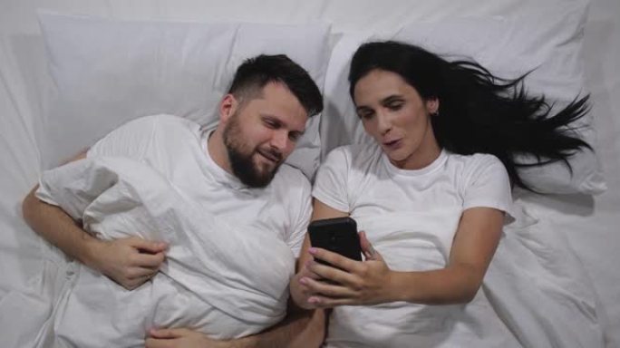 白种人夫妇躺在光明室的床上清醒的俯视图。高角度拍摄。男人和女人在手机上的互联网上看有趣的视频。社交媒