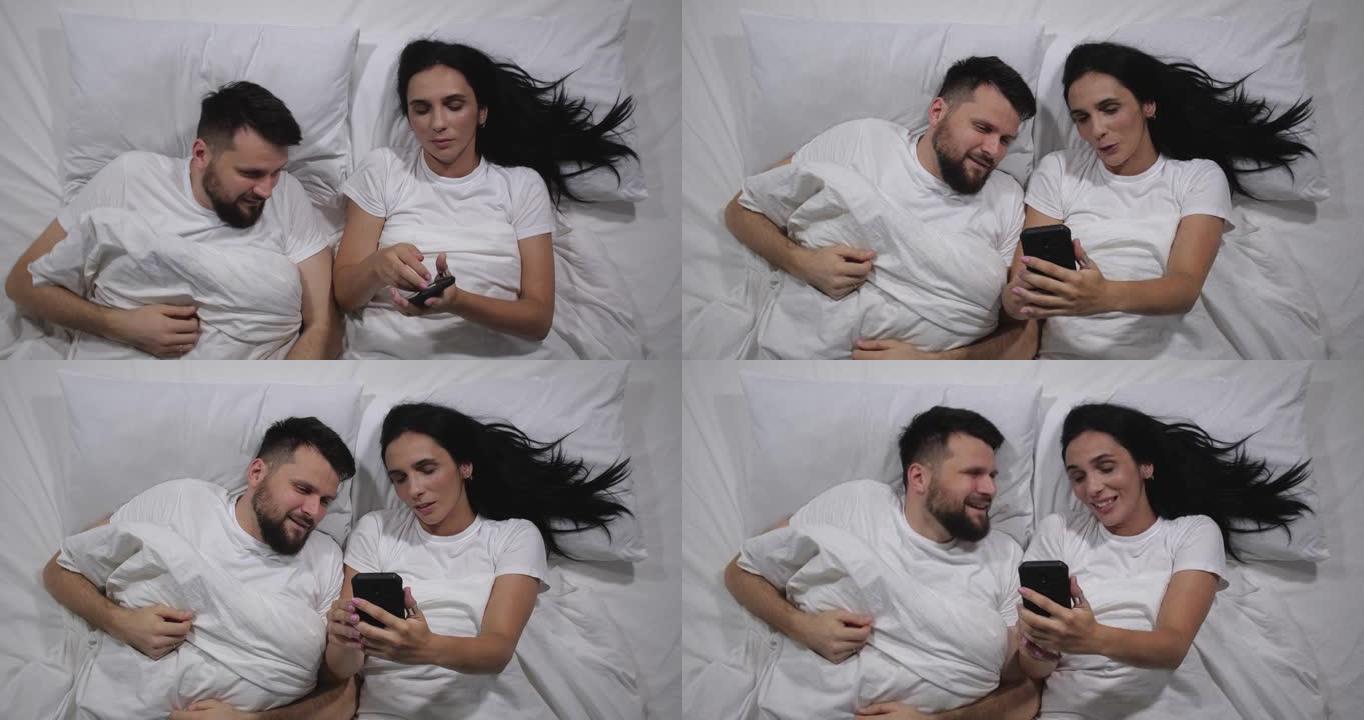 白种人夫妇躺在光明室的床上清醒的俯视图。高角度拍摄。男人和女人在手机上的互联网上看有趣的视频。社交媒