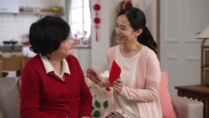 当他们坐在家里的客厅沙发上聊天时，穿着红色衣服的慈爱的亚洲老母亲给儿媳一个红包