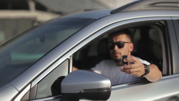 这个人坐在驾驶座上的车里。他正在看摄像机，并展示汽车钥匙。关注关键。他戴着墨镜。4K
