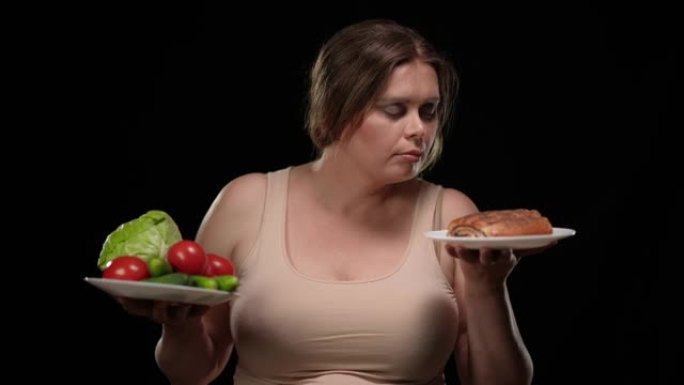 超重妇女在黑色背景下选择不健康的甜甜点与健康蔬菜的肖像。美丽的高加索加大码女士闻到美味的面包微笑。节