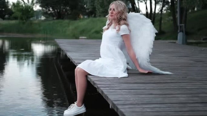 迷人的金发女郎，白色天使翅膀站在水边。穿着天使服装的美女。女神。慢动作