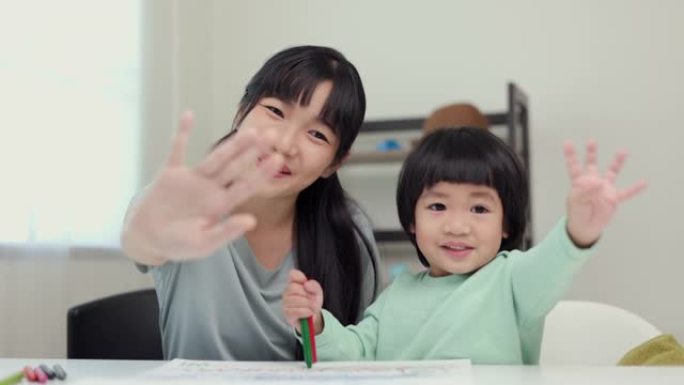 快乐时刻亚洲母亲和小男孩在家里用蜡笔画画时对相机打招呼或再见。家人在房子里玩得开心。妈妈教儿子作业。