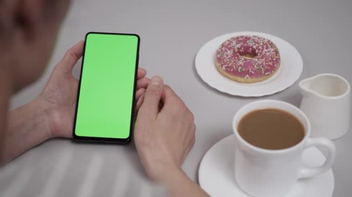 女人坐在餐桌上的咖啡甜甜圈使用带有色度键绿屏的智能手机，滚动浏览社交网络媒体在线商店互联网。智能手机