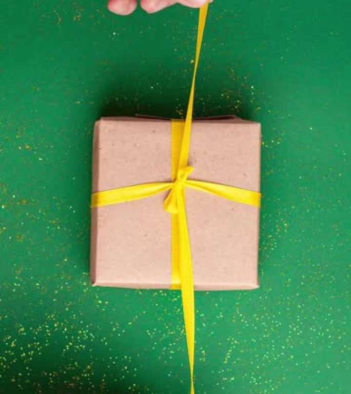 人类双手绑住并解开棕色礼品盒，玉绿色背景上有黄色缎带蝴蝶结，金色闪光。停止运动垂直动画圣诞假期和呈现