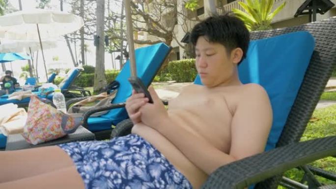 快乐的亚洲男孩使用智能手机，在酒店度假期间放松附近的游泳池，与家人、生活方式理念。
